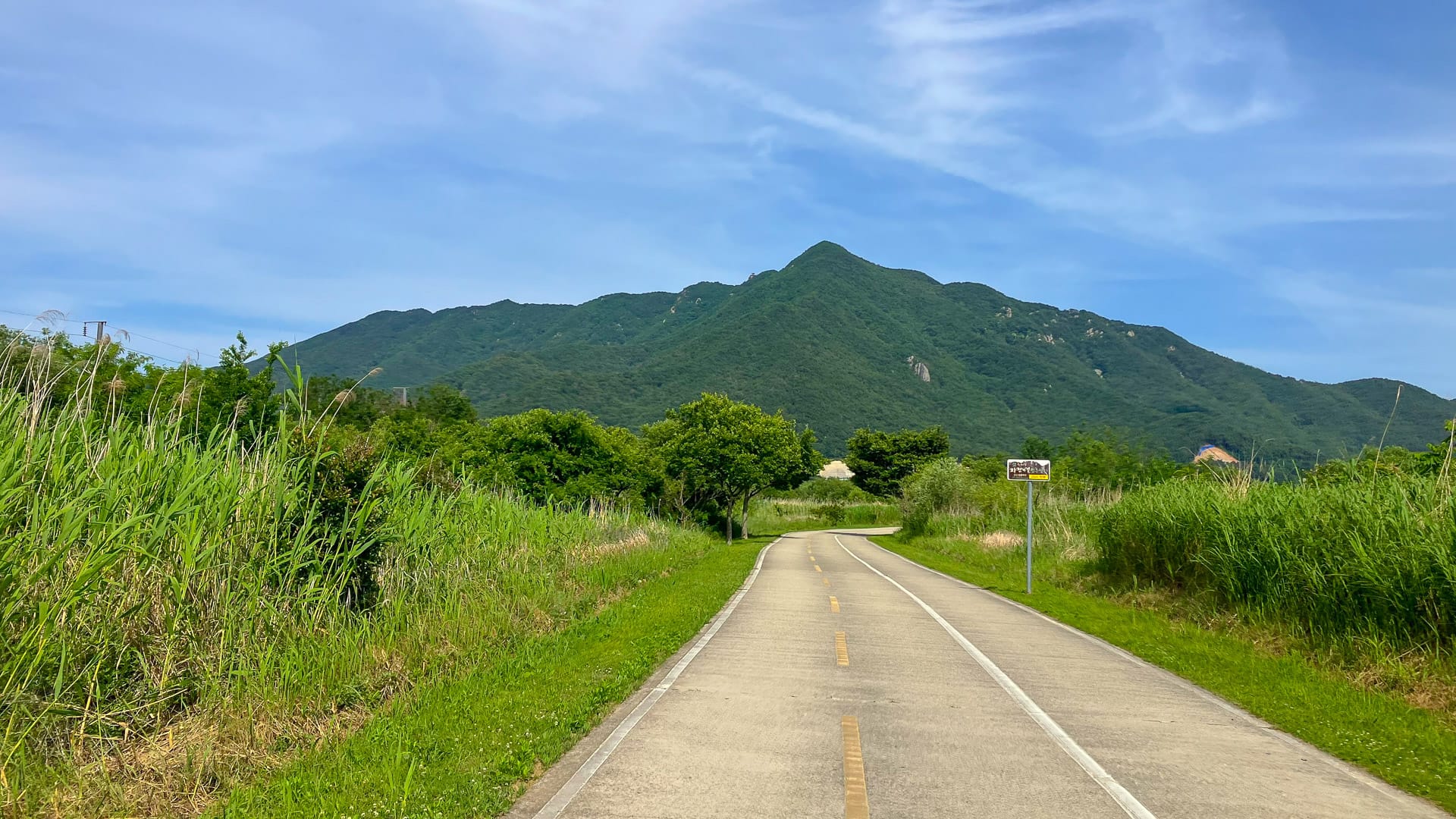 Paved cycle path leading towards a green mountain under a blue sky in rural Korea