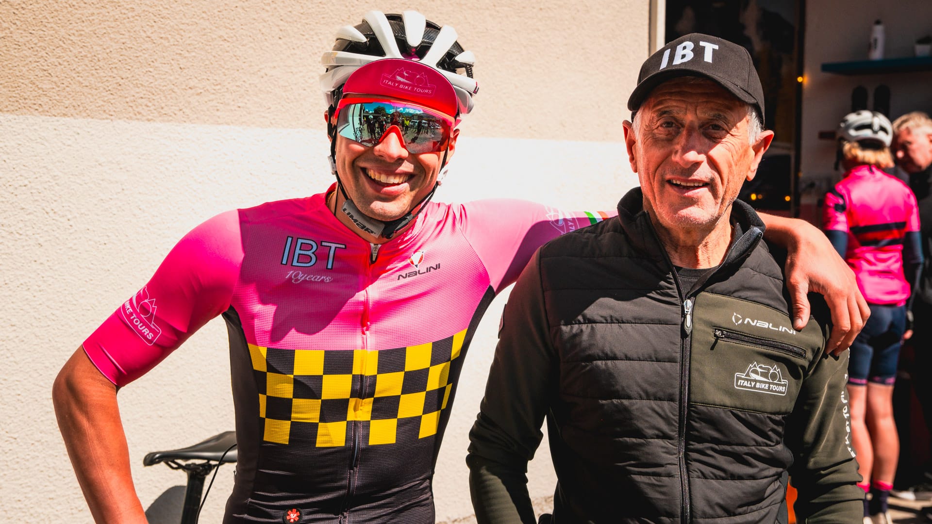 Father and son smiling together, one in a pink cycling jersey and helmet, the other in a black jacket and cap