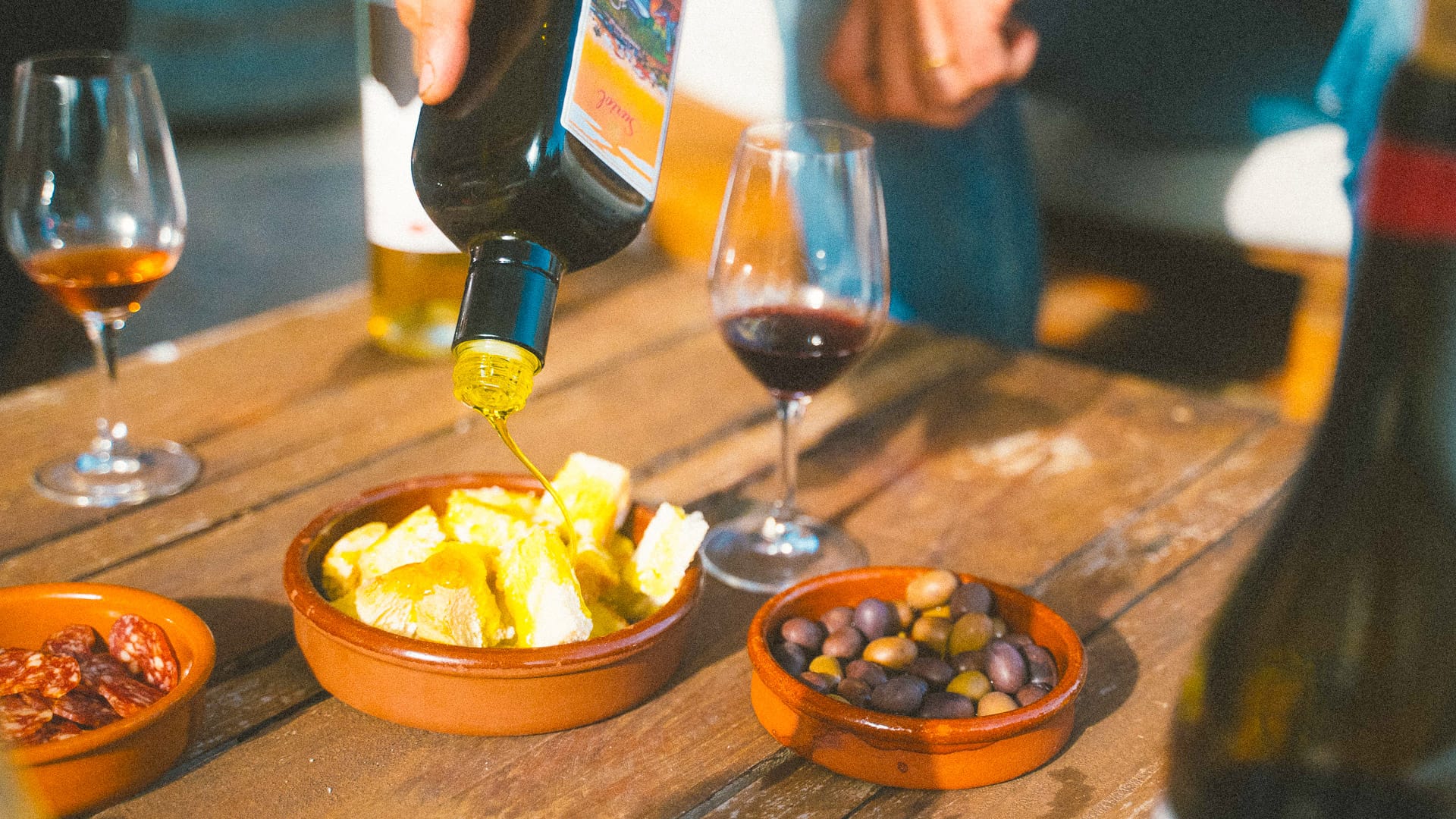Olive oil poured over tapas with olives, potatoes and wine glasses on a rustic wooden table