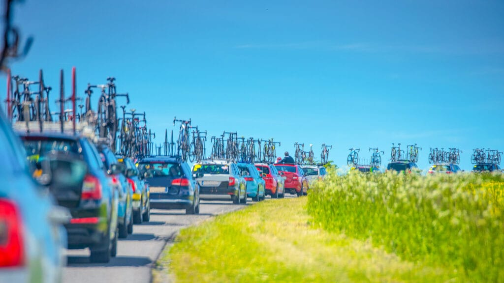 A line of cars carrying bicycles on their roofs drives along a rural road bordered by green fields under a clear blue sky