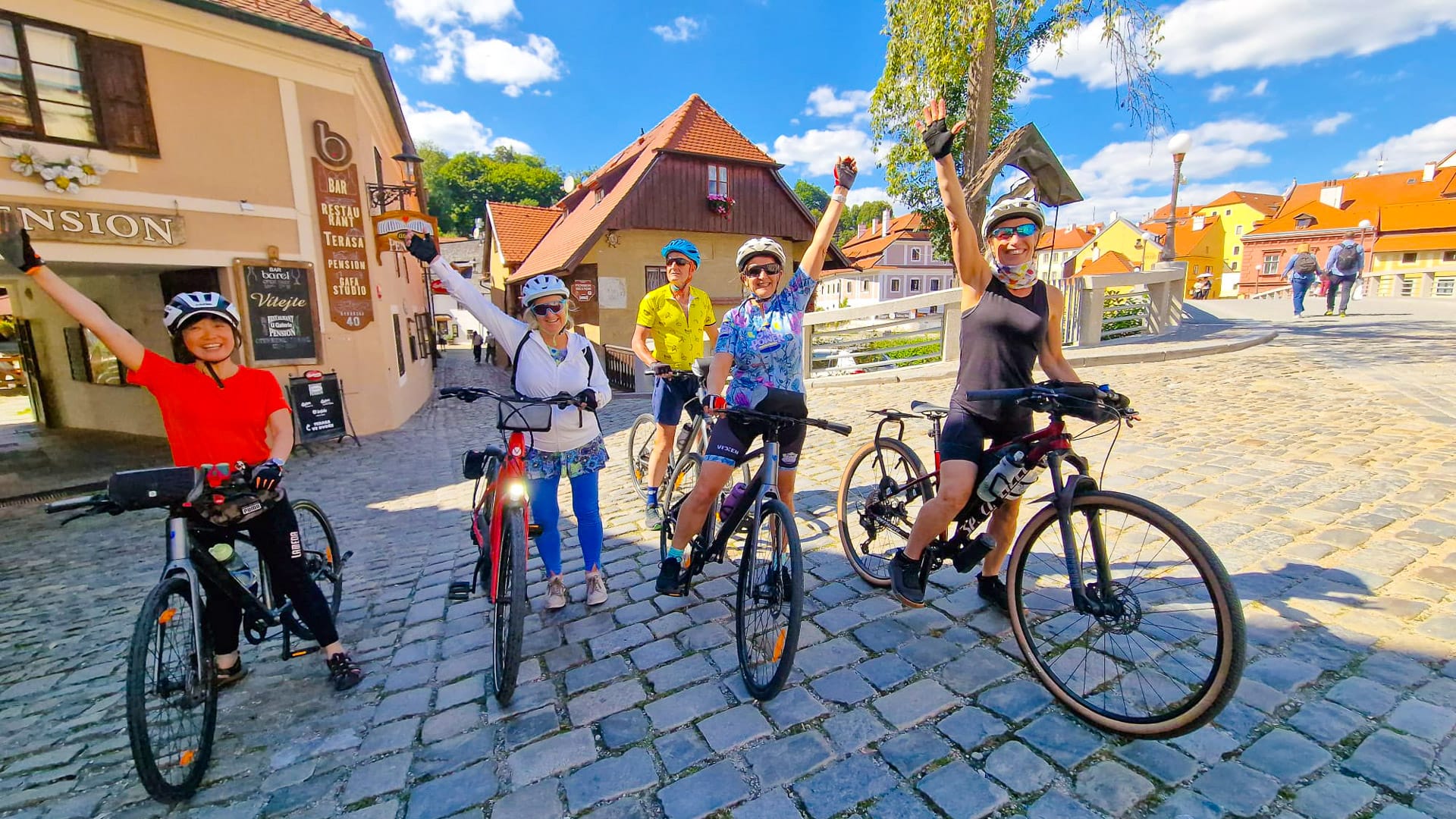 Cyclists raising arms in celebration on a cobbled street surrounded by historic houses in Český Krumlov, Czech Republic