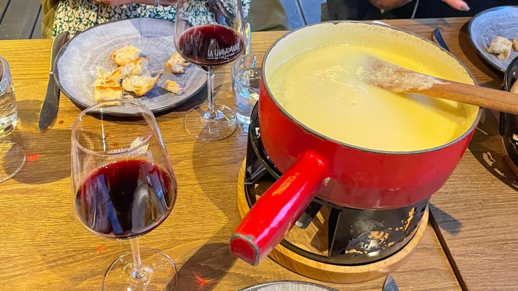 Traditional cheese fondue in a red pot with bread and glasses of red wine on a wooden table
