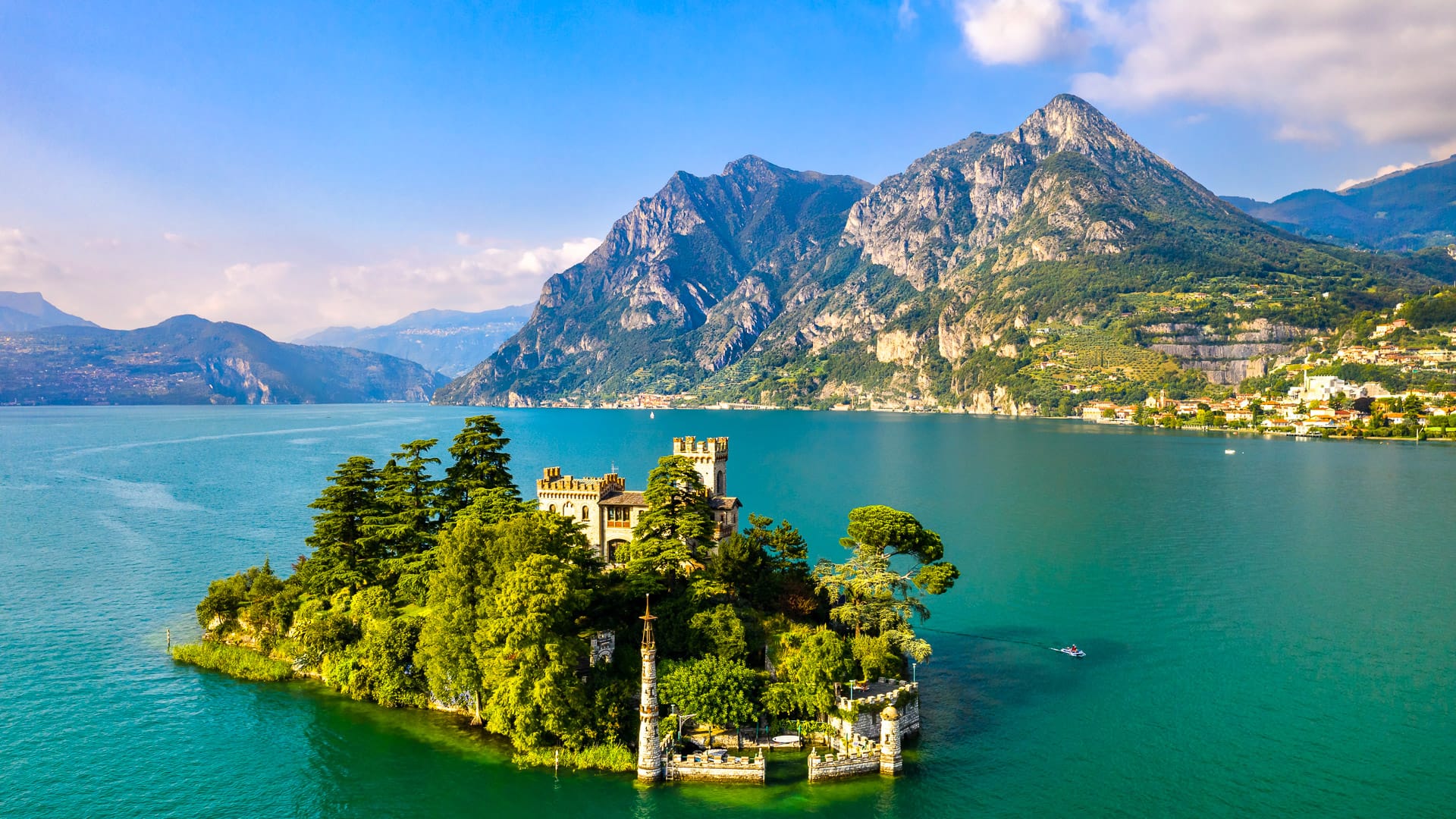 Aerial view of Isola di Loreto, a small island with a castle-like villa and trees, surrounded by Lake Iseo’s turquoise waters and mountains