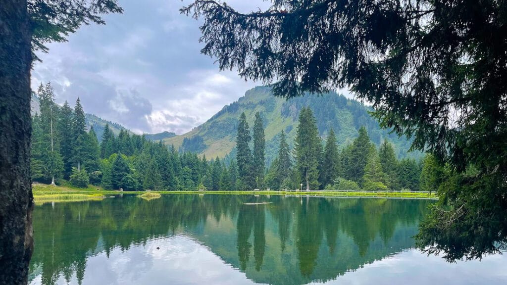 Still waters of Lac de Mines d’Or reflecting surrounding trees and alpine peaks framed by forest branches