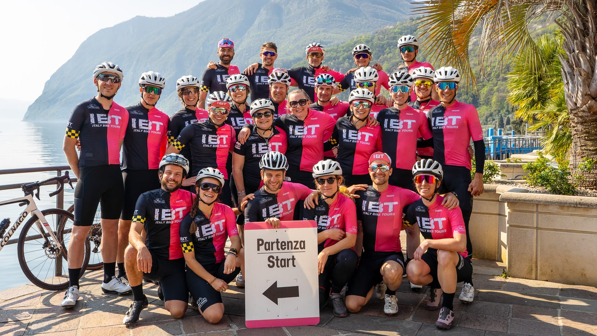 Group of cyclists posing at the start point near a scenic lake with mountains in the background