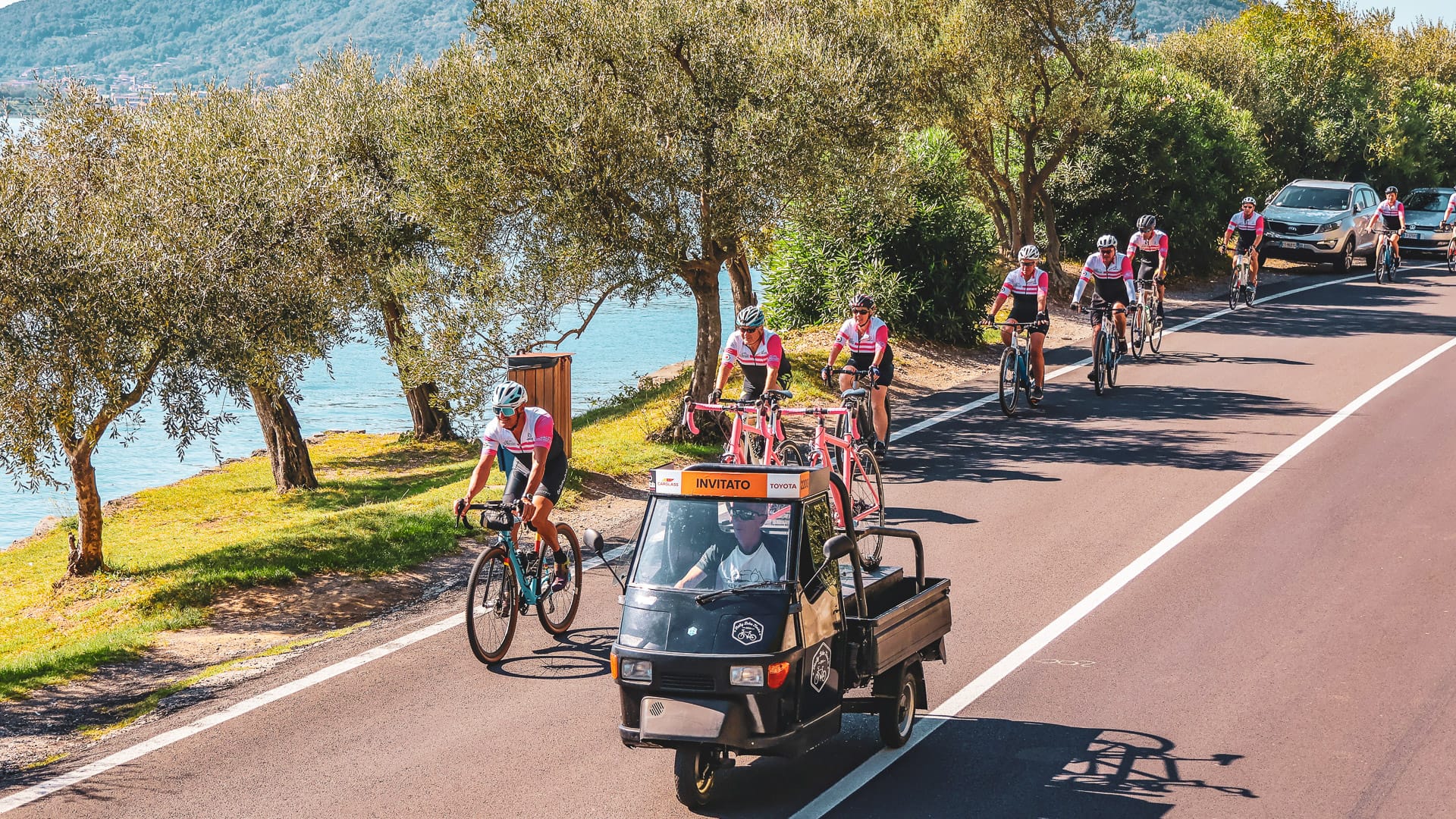 Cycling group following a support vehicle along a sunny lakeside road lined with trees