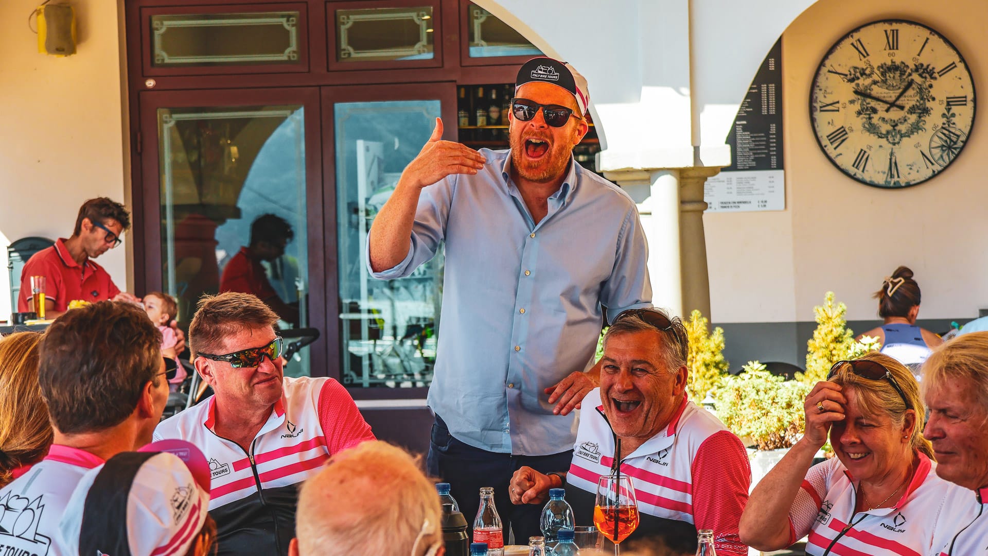 Group of cyclists laughing as a man enthusiastically speaks at an outdoor café in Italy