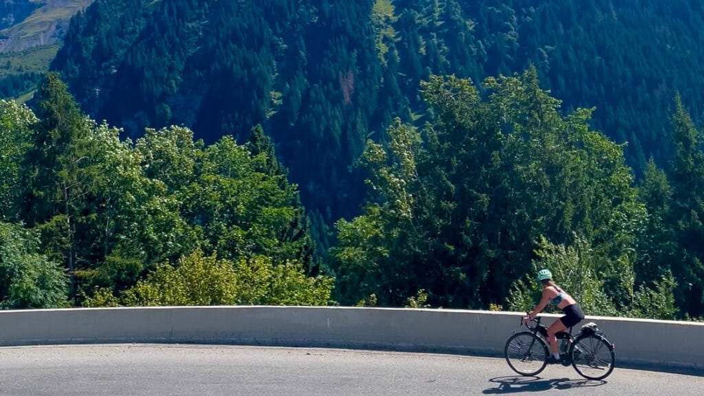 Cyclist rounding a mountain hairpin bend with lush green forest and dramatic alpine backdrop near Pointe de Nyon