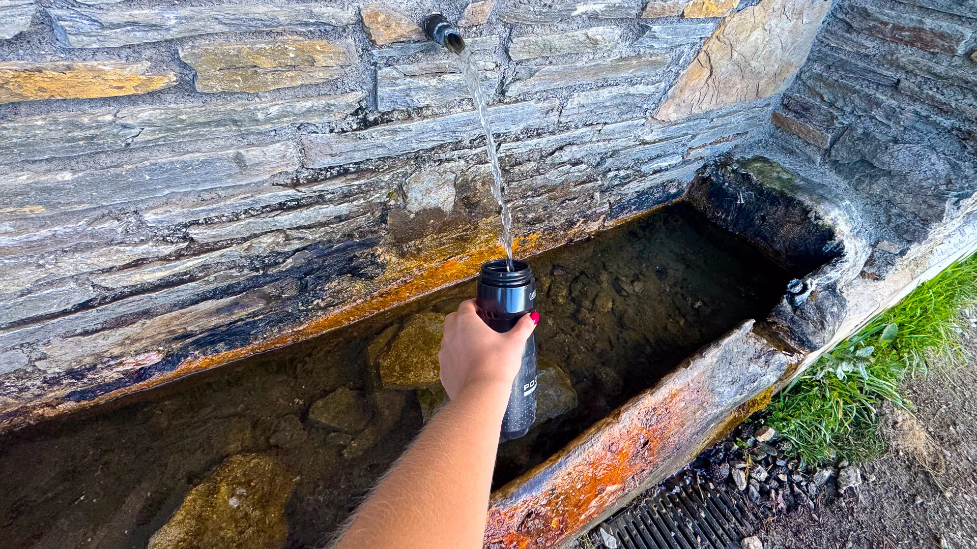 Cyclist refilling bottle from stone fountain at Font del Bosc, Andorra with clear spring water running into a stone trough