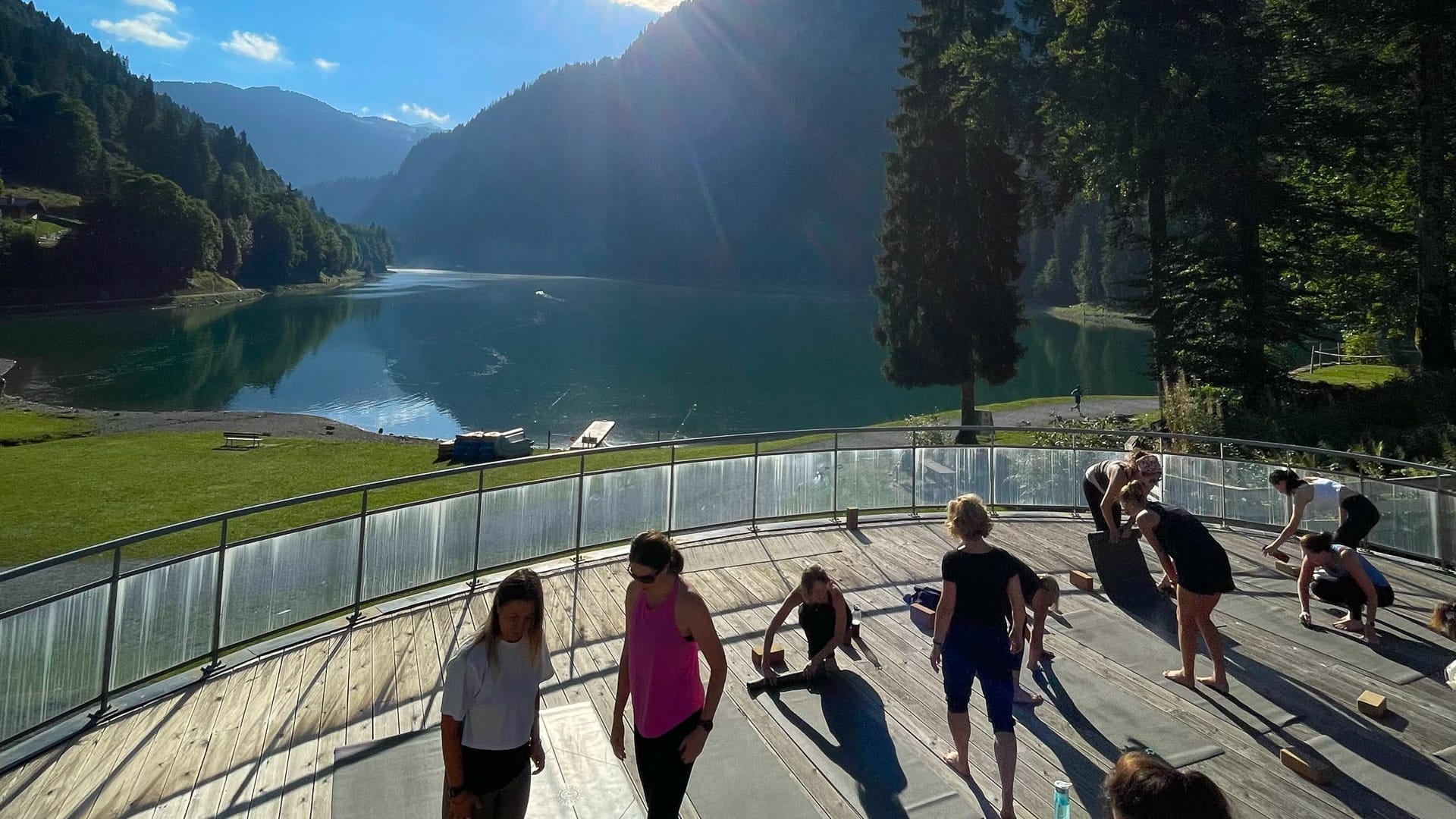 Group doing yoga on a lakeside platform surrounded by forested mountains and morning sunlight streaming across the lake