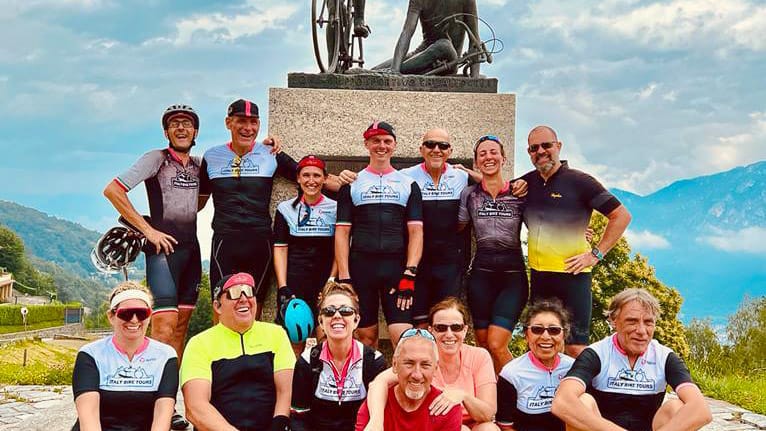 Group of cyclists posing in front of a cycling monument with mountains in the background on a cloudy day