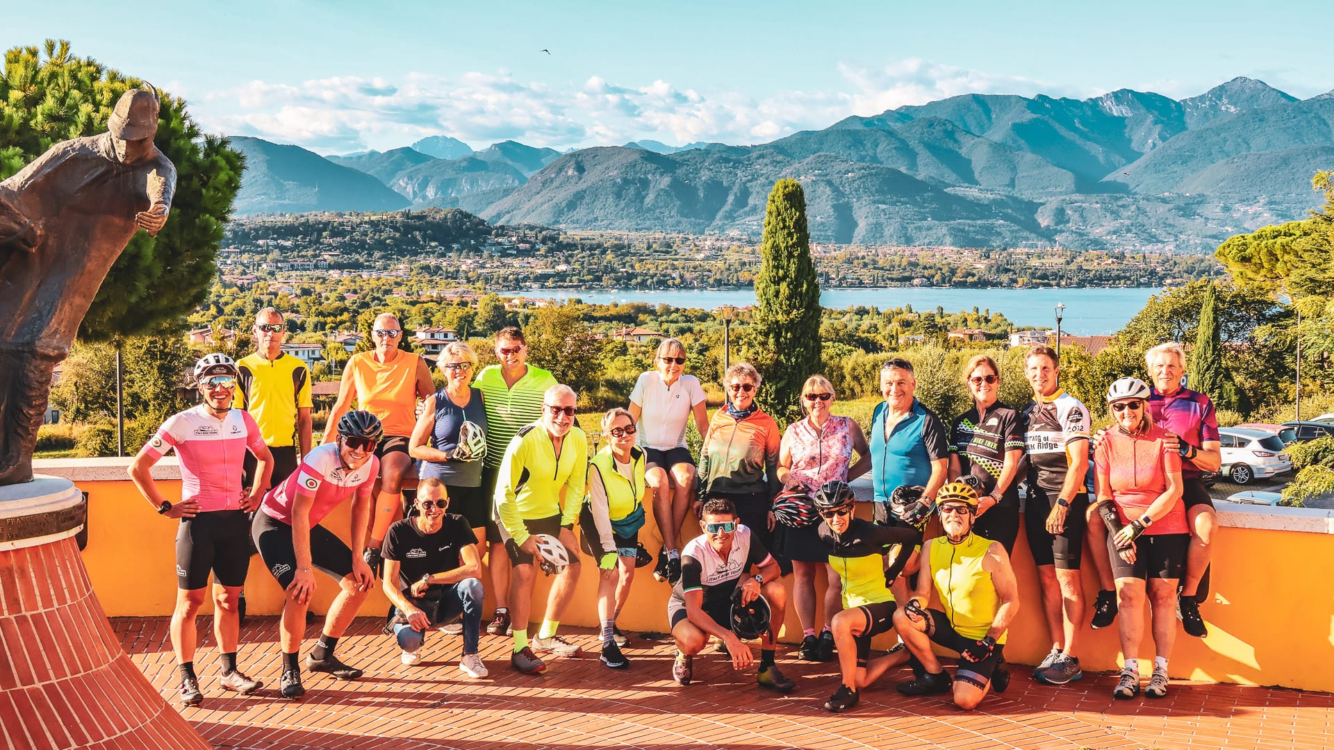 Group of cyclists posing together on a sunny terrace overlooking Lake Garda and surrounding mountains