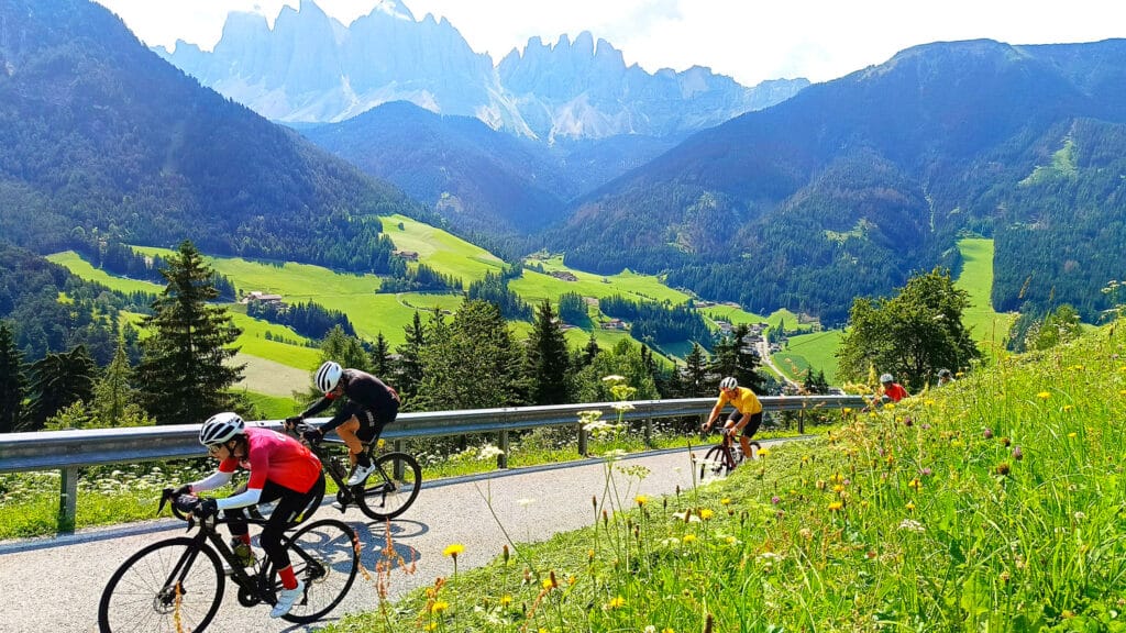 Cyclists climbing a scenic mountain road at Passo delle Erbe wearing colourful jerseys with Dolomites views