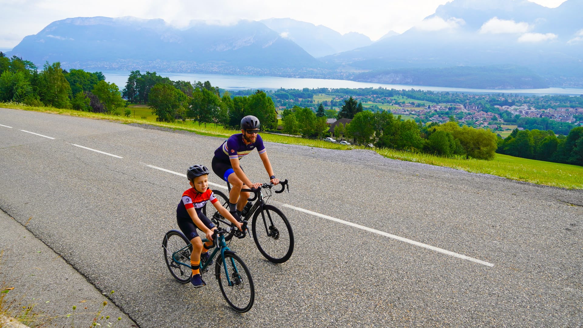 Adult and child cycling uphill with Lake Annecy and alpine peaks in the background, French Alps
