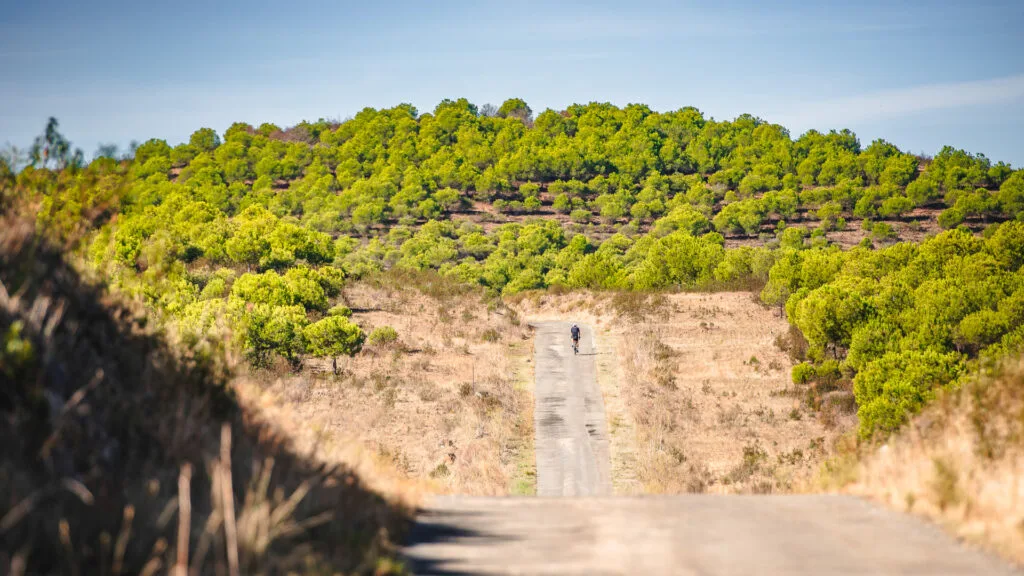 Cyclist riding up a quiet rural road lined with green hills during the BikingMan Portugal ultracycling race