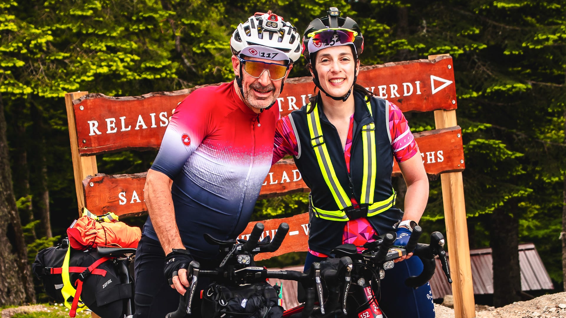 Two cyclists smiling with their loaded bikes at a mountain signpost during BikingMan Corsica