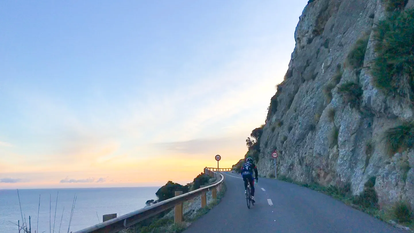 Cyclist climbing a coastal cliff road at sunset above the Mediterranean near Cap de Formentor, Mallorca