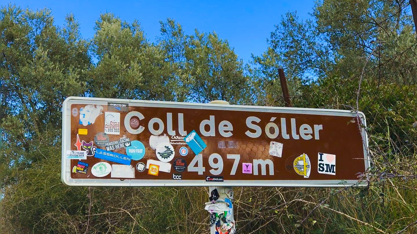 Brown Coll de Sóller summit sign at 497 m in Mallorca, covered in stickers, with trees and clear sky behind