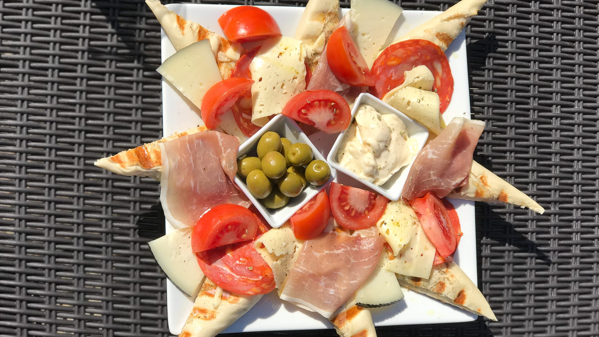Spanish tapas plate with cheese, tomato, ham, olives and dips served outdoors on woven surface under strong midday sun