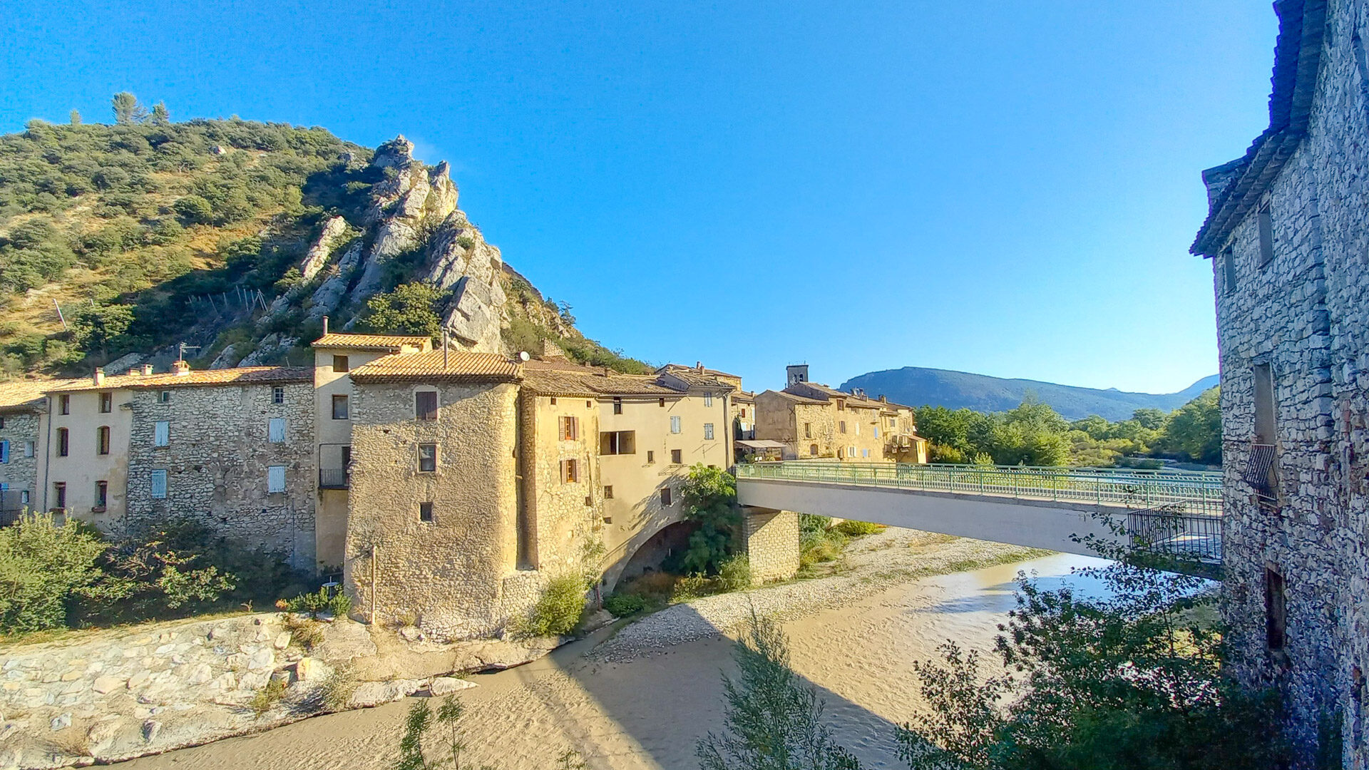 Sunny view of an old stone village with a bridge over a shallow river, set against rocky hills and a clear blue sky