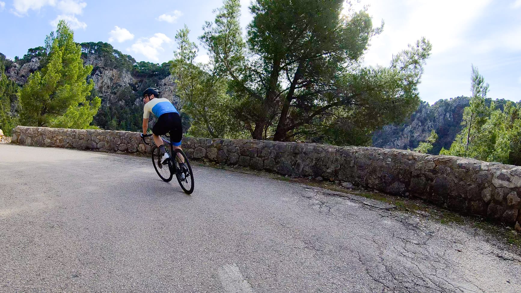Cyclist rounding a bend on a quiet descent above Port de Valldemossa, Mallorca, surrounded by stone wall and pine forest