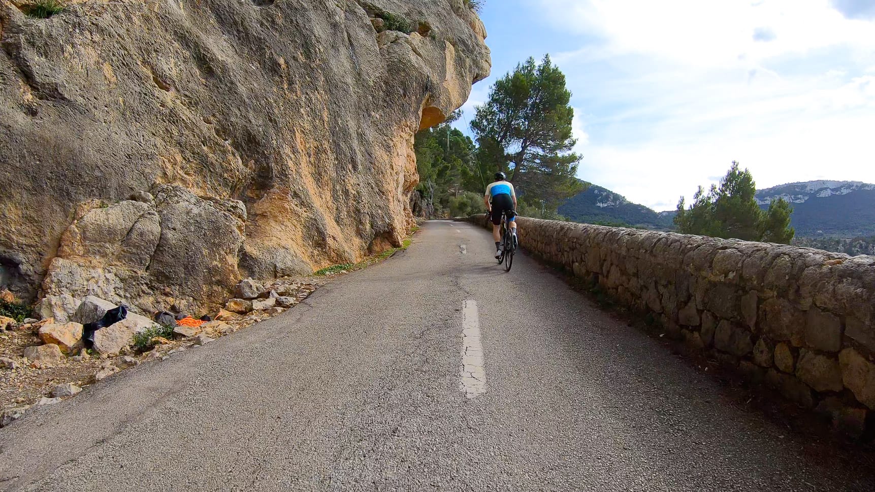 Cyclist riding uphill past a rock overhang on Mallorca’s Port de Valldemossa road, lined with pine trees and low stone barrier