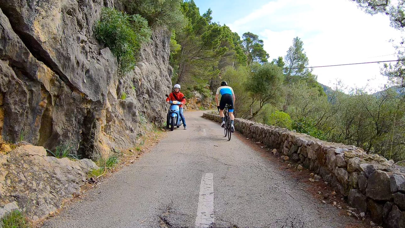 Cyclist passing parked scooter on narrow cliffside road in Mallorca, lined with stone wall and pine trees