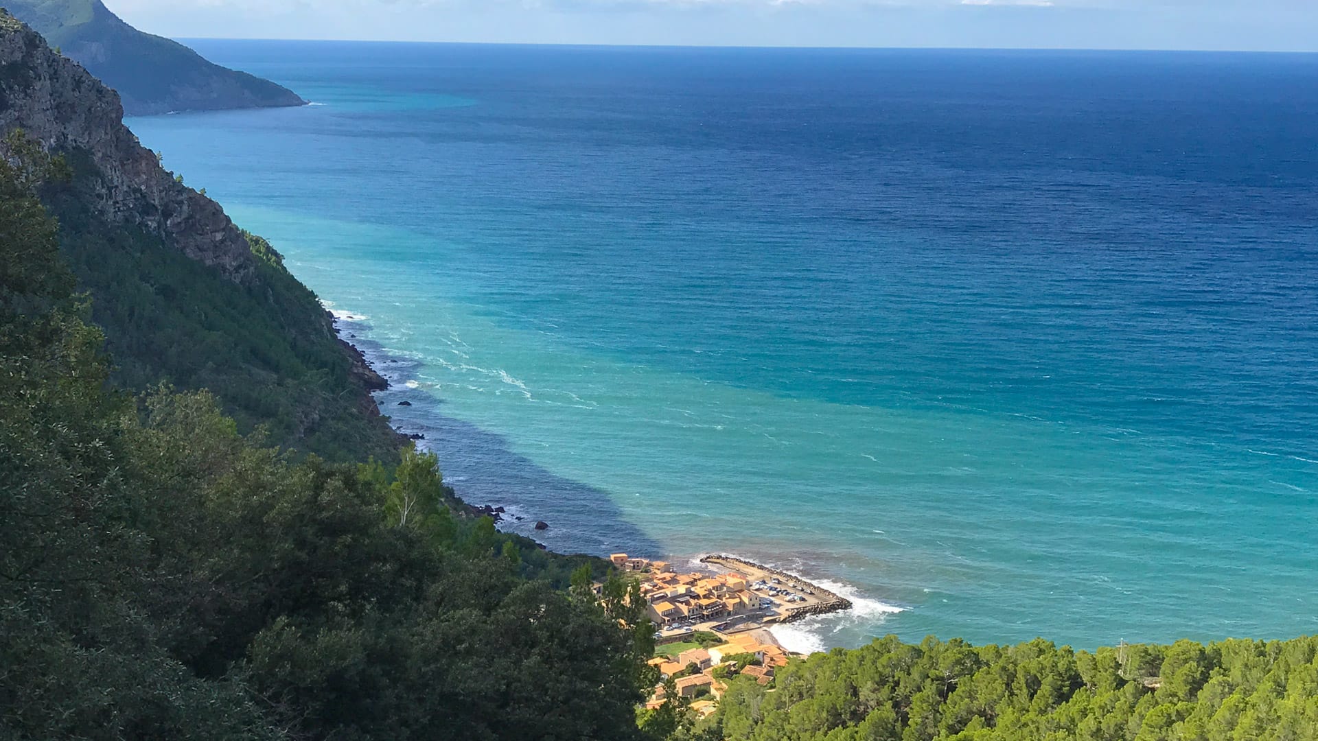 View of Port de Valldemossa, Mallorca from above, with turquoise sea, forested cliffs and winding road leading down to the coast