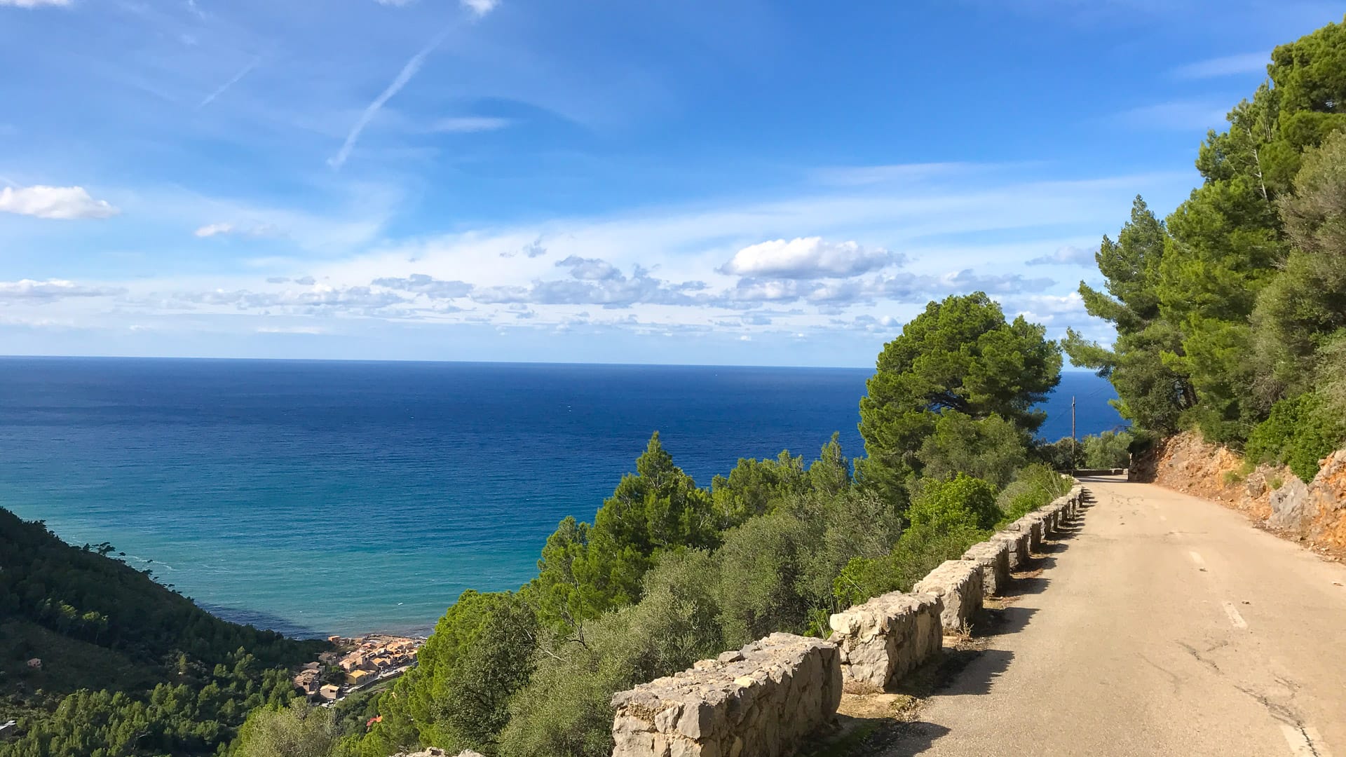 Quiet coastal descent road above Port de Valldemossa, Mallorca, with sea views and stone barriers along tree-lined bends