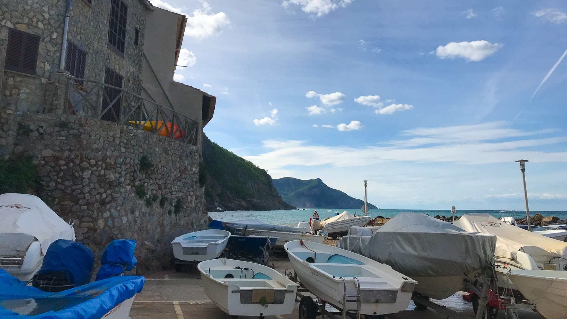 Boats and dinghies lined up beside stone buildings at Port de Valldemossa, Mallorca, with coastal cliffs and calm sea behind