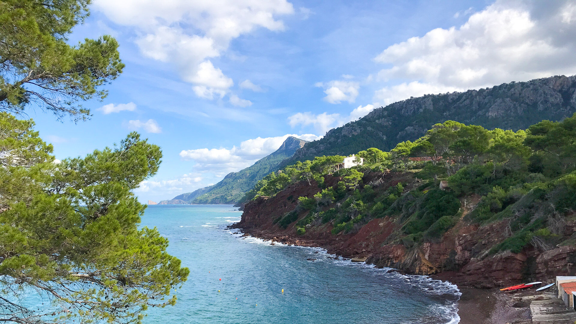 Coastal cove on Mallorca’s northwest shore with red cliffs, pine trees and calm turquoise sea beneath a partly cloudy sky