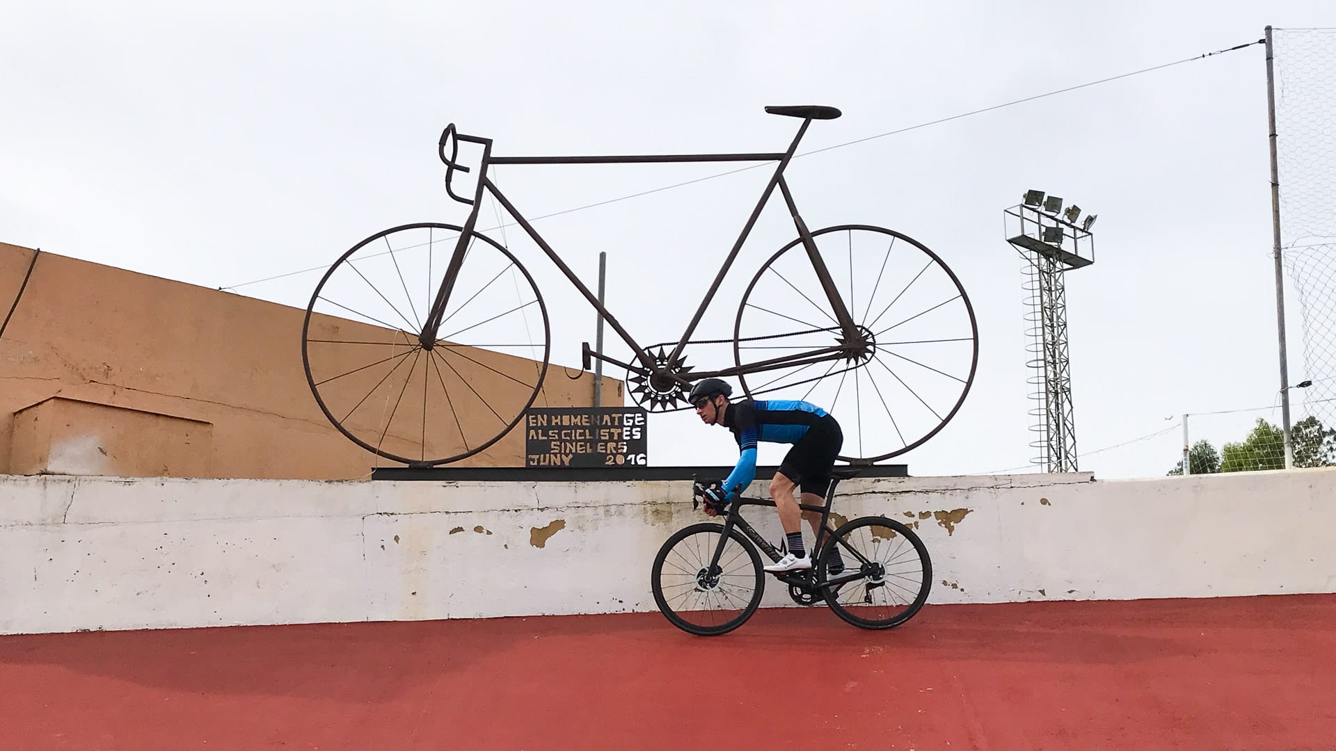 Rider on the red banked curve at Sineu Velodrome beneath oversized steel sculpture of a penny-farthing bicycle