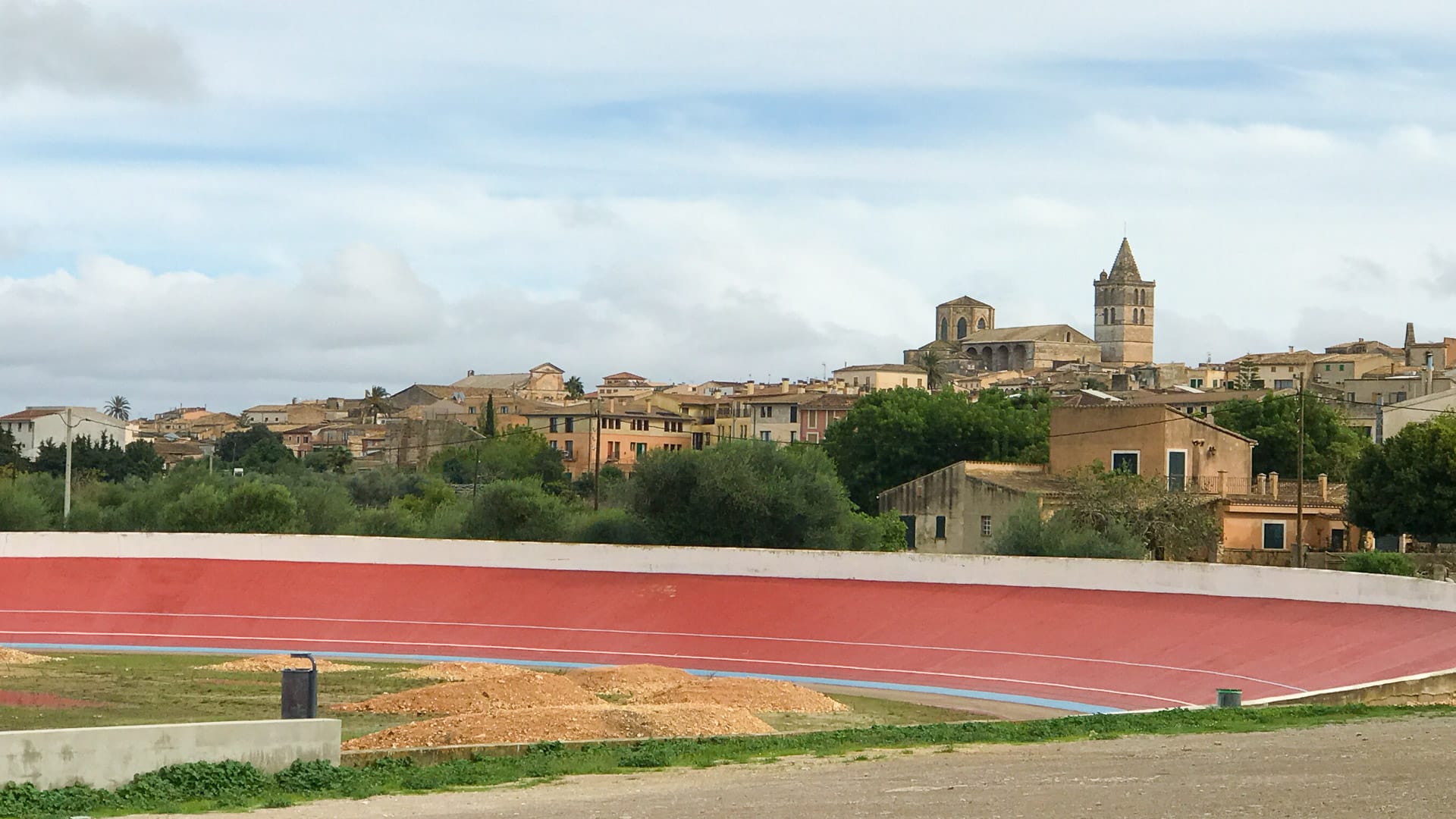 View of Sineu town and church tower rising above tan rooftops, with the red curve of the velodrome track in the foreground