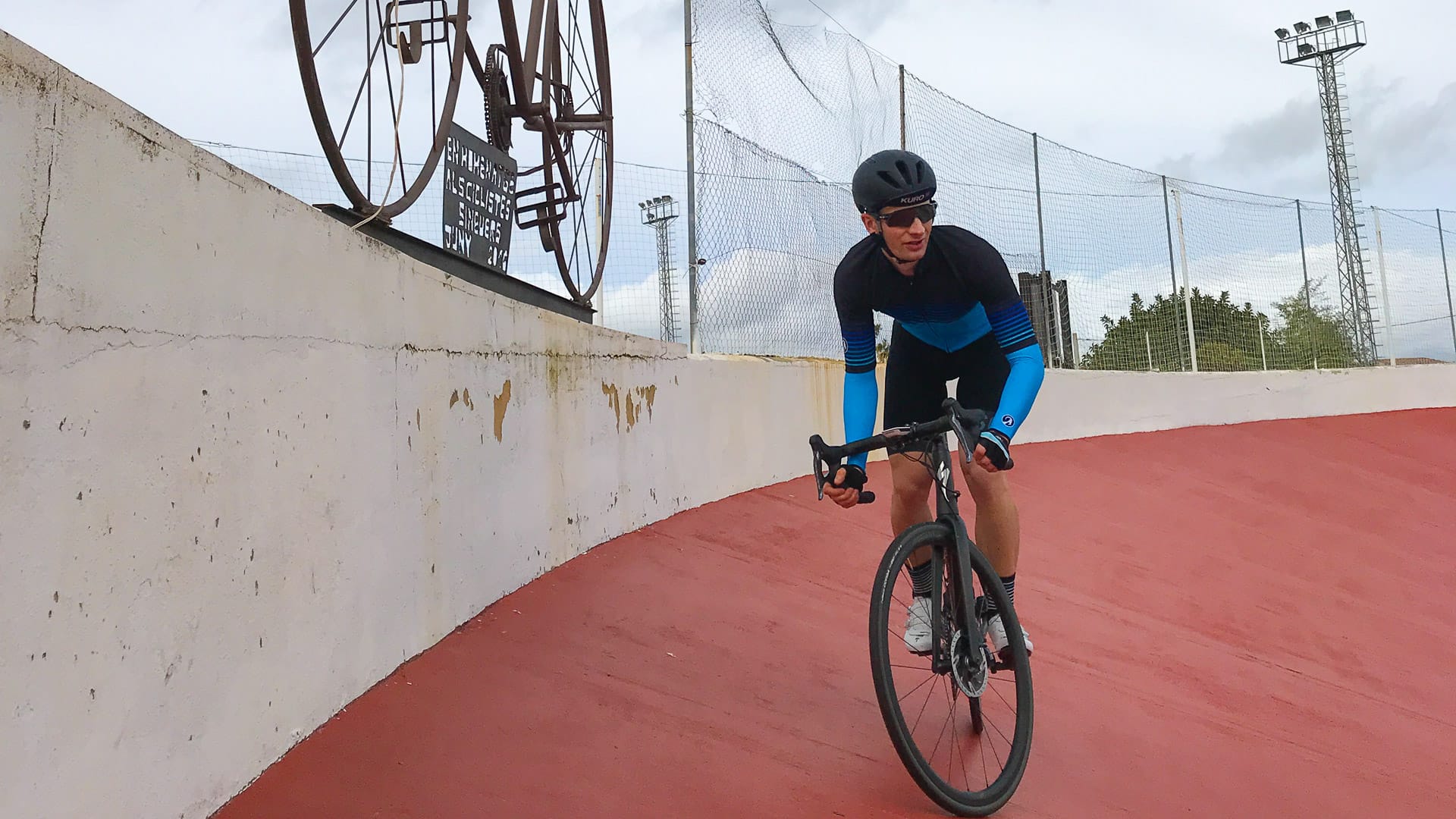 Cyclist in black and blue kit riding on steep red banking at Sineu velodrome, Mallorca, with vintage bicycle sculpture above wall