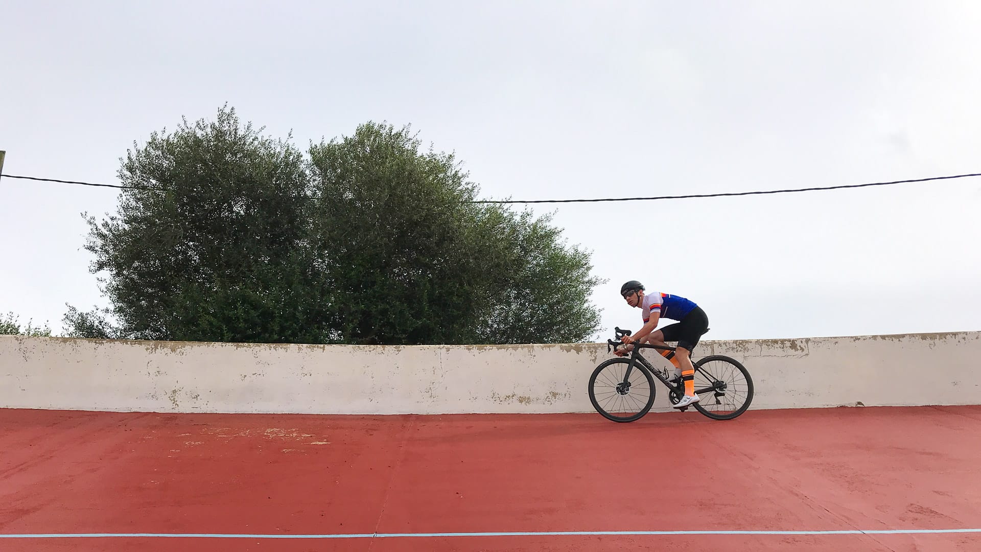 Cyclist on red track surface at Sineu velodrome, Mallorca, riding past low outer wall and tree in the background