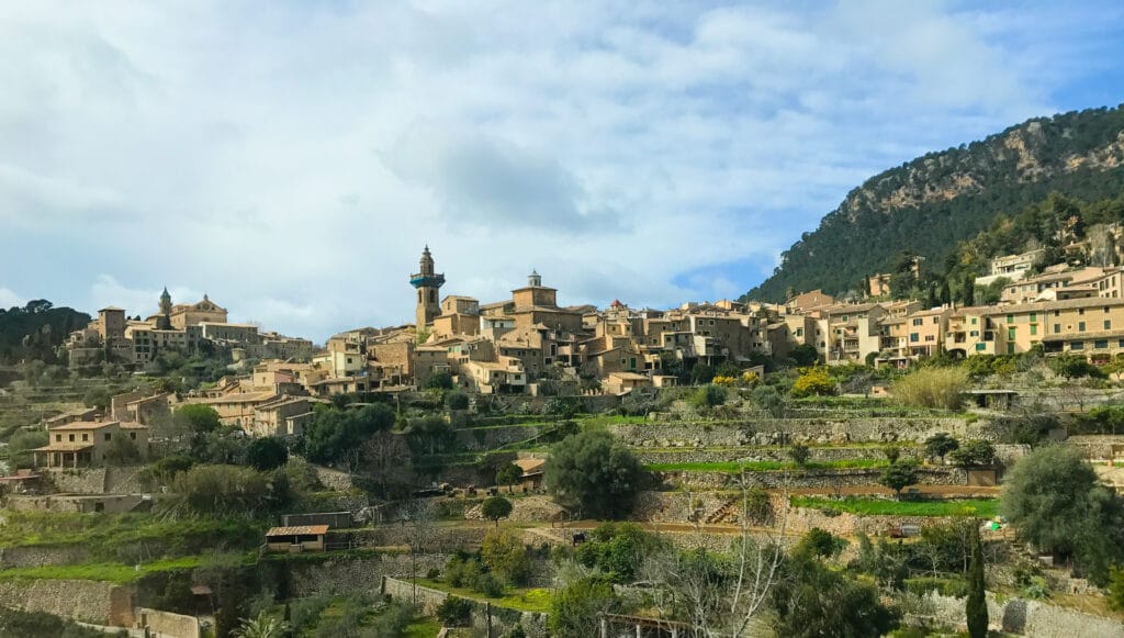Hilltop village of Deià, Mallorca with terraced fields, stone houses and church tower nestled against forested slopes under a bright sky