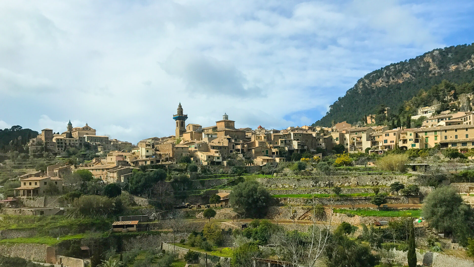 Hilltop village of Deià, Mallorca with terraced fields, stone houses and church tower nestled against forested slopes under a bright sky
