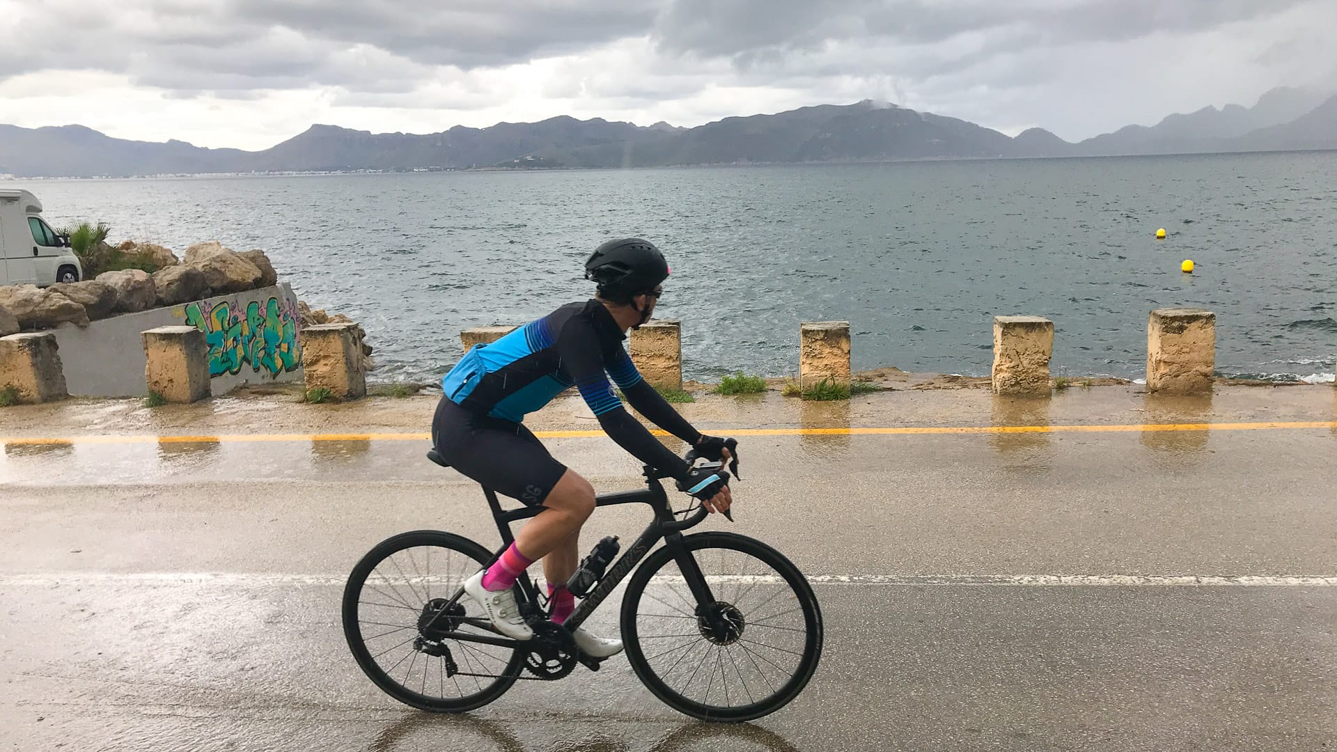 Cyclist riding along wet seafront road in the rain with bay and mountains across Pollenca bay
