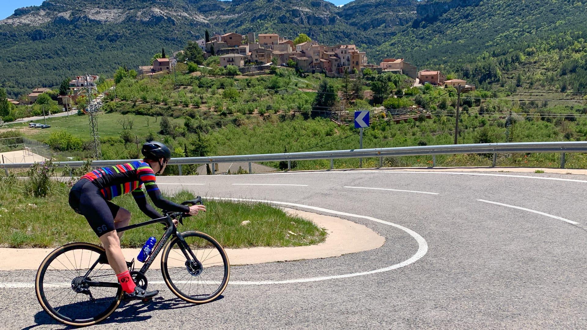 Cyclist riding a mountain road with a hilltop village and rugged cliffs in the distance on a sunny day