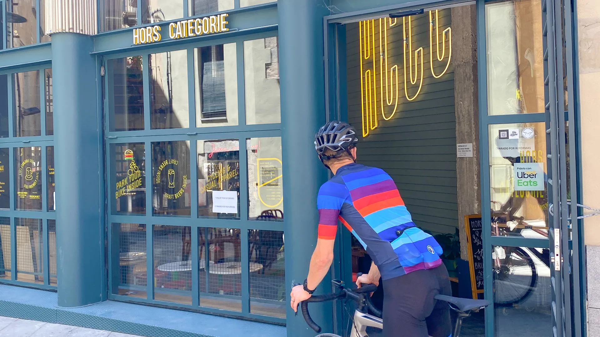 Cyclist in rainbow jersey entering Hors Catégorie café in Girona through glass-fronted entrance
