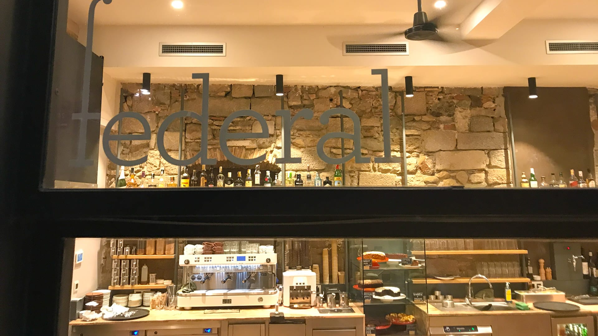 View through window of Federal Café in Girona showing espresso machine, cakes and bottles behind stone wall interior