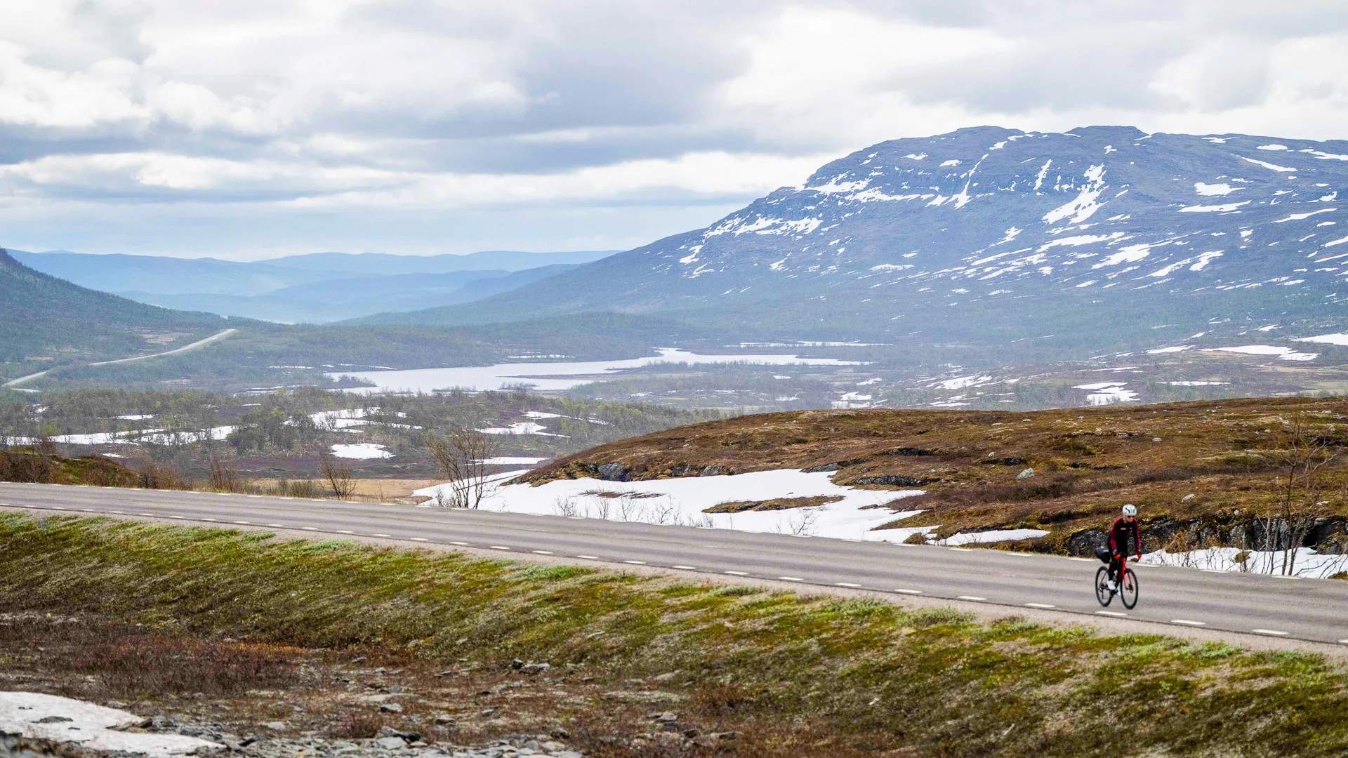 Solo cyclist on a remote road with snow patches and mountains during MSR 2025 in northern Sweden