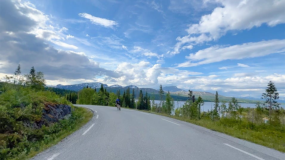 Cyclist on a quiet lakeside road in northern Sweden with forest and mountains under a bright sky