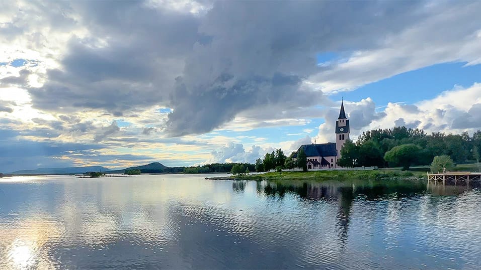 Lakeside church in Västerbotten Sweden with tall steeple reflected in calm water under dramatic clouds