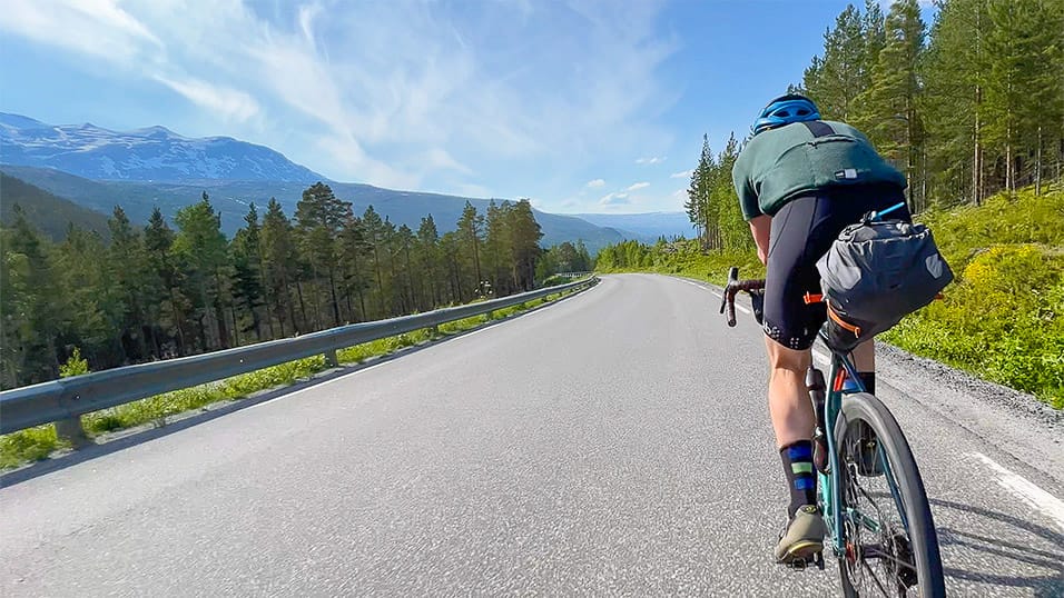Cyclist climbing a sunny mountain road through pine forest with distant snow-capped peaks in northern Sweden