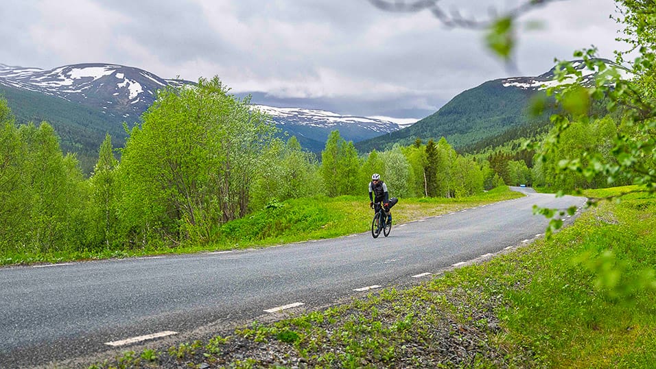 Solo cyclist riding a quiet mountain road near snow-topped peaks in northern Sweden