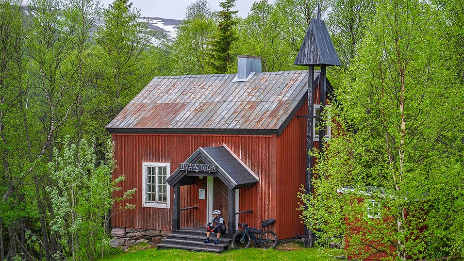 Cyclist resting outside a small red wooden chapel surrounded by spring forest near Hemavan Sweden