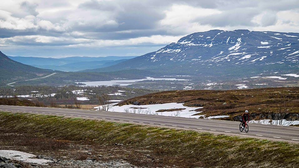 Cyclist riding across an open mountain plateau in northern Sweden with snow patches and distant lakes