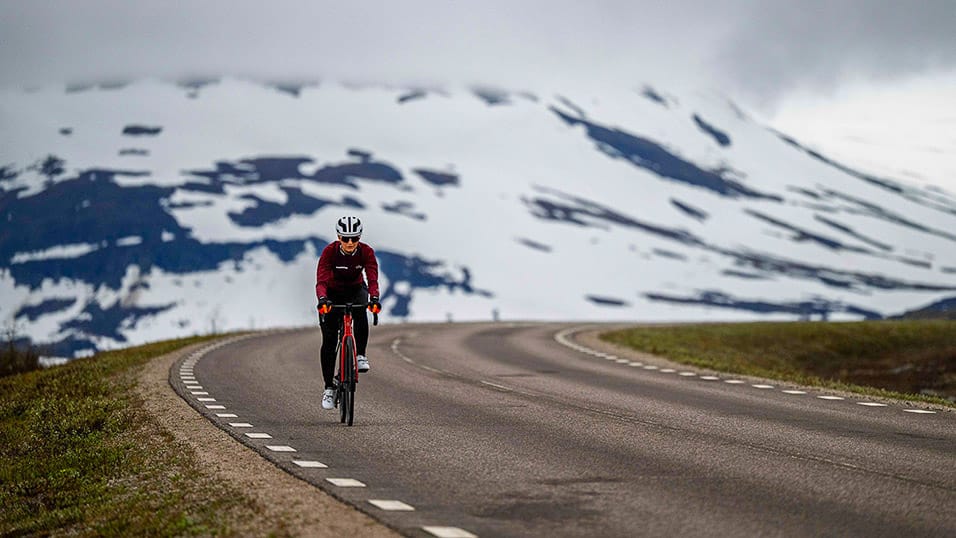 Cyclist riding along a winding road beneath snow-covered mountains during the Midnight Sun Randonnée in Sweden