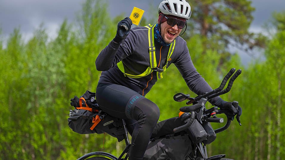 Smiling rider holding up a yellow brevet card while cycling the Midnight Sun Randonnée in Västerbotten Sweden